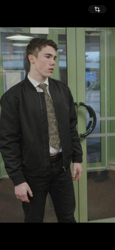 Young man in a black bomber jacket and tie, standing in an indoor setting.