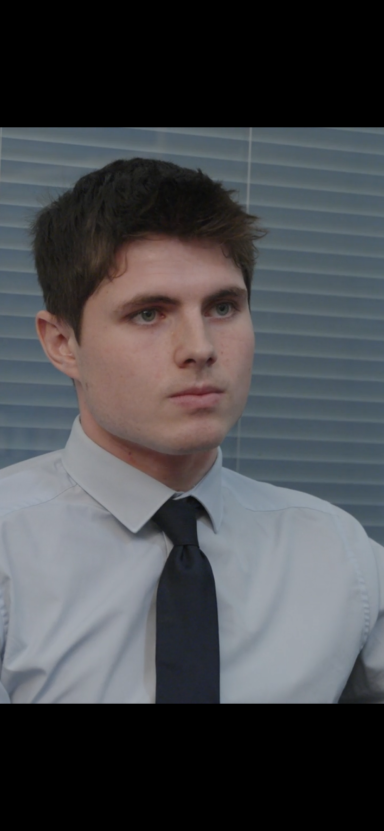 Young man in a light grey shirt and dark tie, looking serious in an office setting.