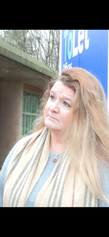 Woman with long hair, wearing a scarf, standing near a toilet sign outdoors.