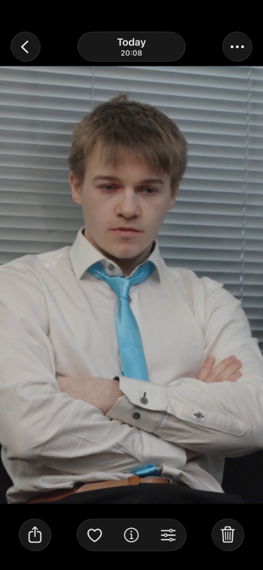 Young man in a white shirt and blue tie sitting with arms crossed, looking serious.