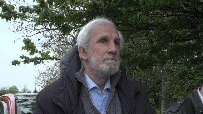 Elderly man with grey hair, wearing a coat, looking contemplatively outdoors.