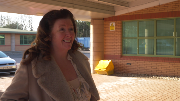 Smiling woman in a light coat stands outside a building with a green window.