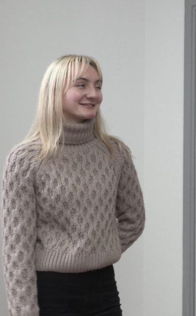 Smiling young woman with blonde hair, wearing a light sweater and black trousers.