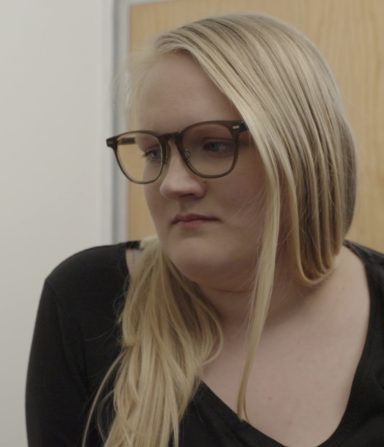 Young woman with long blonde hair and glasses, looking thoughtfully to the side.