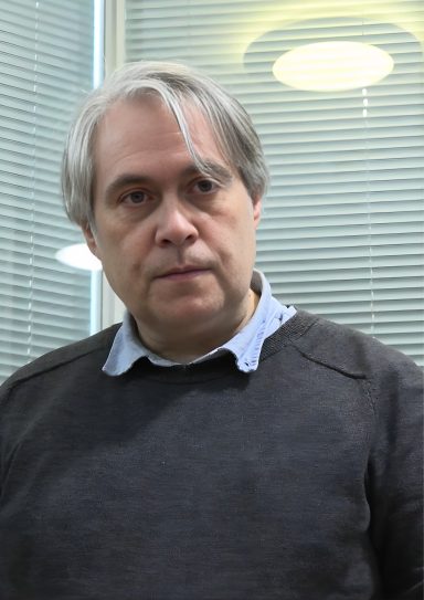 A man with grey hair and a serious expression stands in a well-lit indoor setting.