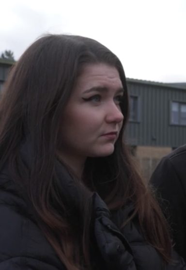 A young woman with long dark hair, wearing a black jacket, appears contemplative outdoors.