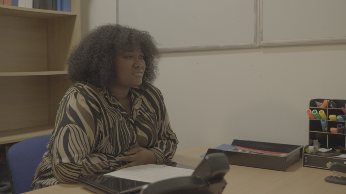 A woman with curly hair sits at a desk, engaged in conversation.